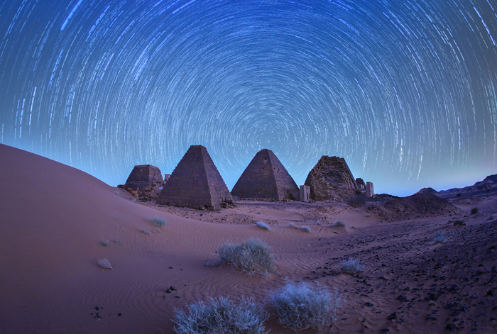 Four pyramids are sitting in the middle of sand hills. The sky is blue with a circular effect created by the movement of the earth over time.